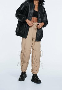 Veste aviateur noire en similicuir, pantalon cargo beige avec cordons de serrage, et bottes à lacets noires épaisses. Tissu texturé et coupe décontractée.