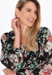 Blusa nera floreale con motivi verdi e rosa; modella che indossa orecchini circolari con base circolare arancione e parte superiore trasparente.