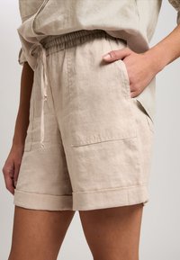 Beige Leinen-Shorts mit elastischem Bund, ausgestattet mit zwei seitlichen Taschen und umgeschlagenen Säumen. Glatte Textur, minimales Design, funktioneller Stil.