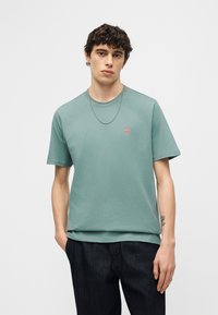 Νεαρός άνδρας με σγουρά σκούρα μαλλιά, φοράει μια απαλό πράσινο κοντομάνικο μπλουζάκι με ένα μικρό στρογγυλό λογότυπο στο στήθος και σκούρο παντελόνι.