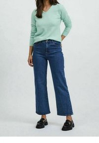 Hellgrüner V-Ausschnitt-Pullover aus weichem Material. Dunkelblaue weit geschnittene Jeans aus Baumwoll-Denim. Schwarze Loafers mit dicken Sohlen. Einfaches, klares Design.