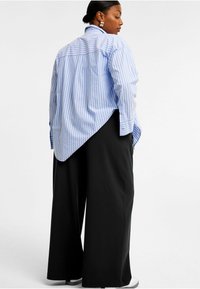 Ljusblå och vitrandig skjorta med hög-låg nederkant, i kombination med lösa svarta byxor. Mjuk tygkvalitet, knäppte cuffs och sidofickor är synliga.