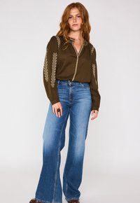 Blusa verde oliva con dettagli ricamati, patta con bottoni e maniche lunghe, abbinata a jeans in denim blu a vita alta e a gamba svasata.