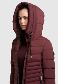 Burgundy puffer jacket dengan tudung tinggi, ritsleting depan, dan aksen hitam. Memiliki bagian berlapis quilted dan tali untuk penyesuaian ukuran.