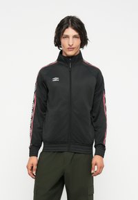 Mlad moški, oblečen v črno zip-up jakno Umbro z rde�čimi in belimi črtami na rokavih ter olivno zelenimi cargo hlačami, stoji pred belo ozadjem.