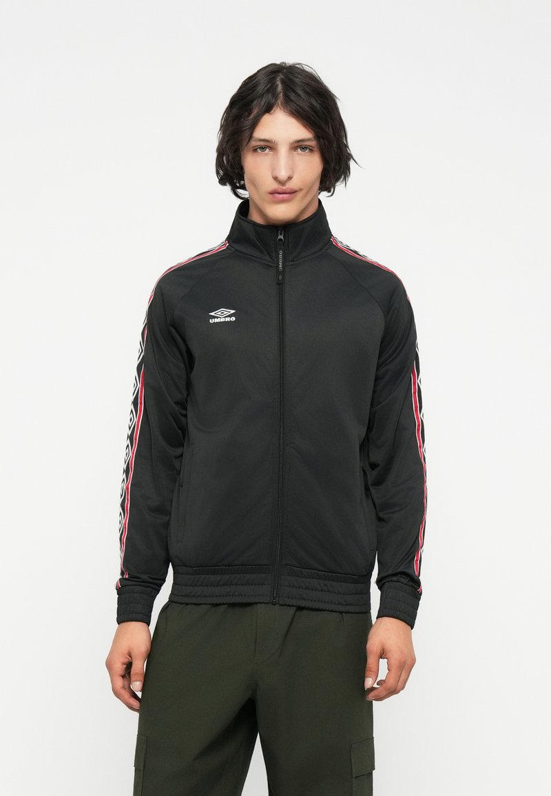 Mlad moški, oblečen v črno zip-up jakno Umbro z rdečimi in belimi črtami na rokavih ter olivno zelenimi cargo hlačami, stoji pred belo ozadjem.