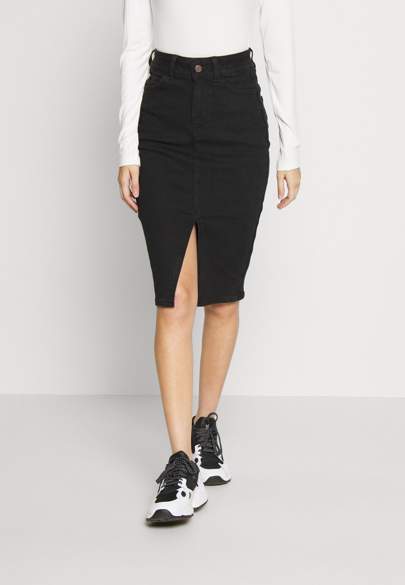 Falda lápiz de mezclilla negra con una abertura frontal, cintura alta y diseño ajustado, combinada con zapatillas negras y blancas. Textura suave, sin estampados.