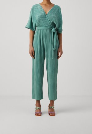 Combinaison verte plissée avec un devant portefeuille et des manches larges, dotée d'une ceinture assortie à la taille. Portée avec des sandales à brides multicolores.