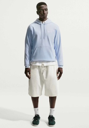 Mężczyzna ubrany w jasnoniebieską bluzę z kapturem, białą koszulę, białe spodenki do kolan, białe skarpetki Nike i ciemnozielone trampki, stojący na jednolitym tle.