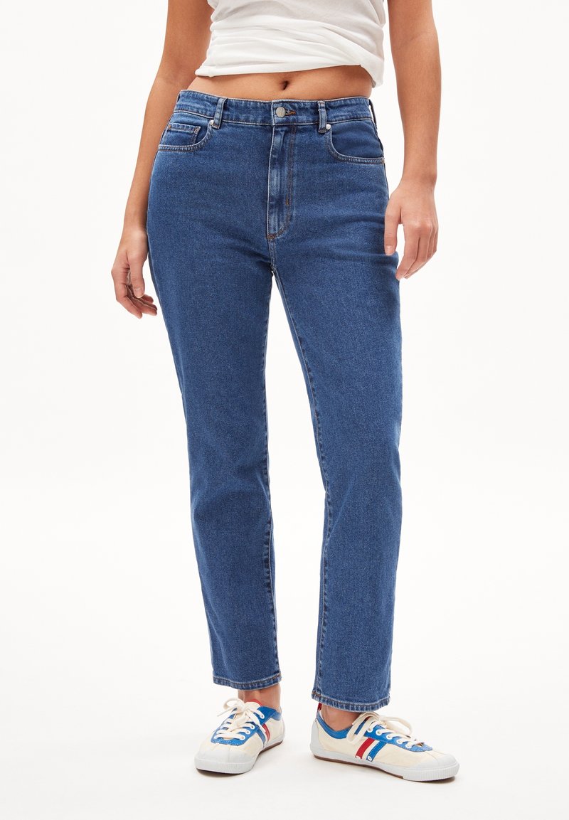 Personne portant un jean bleu taille haute à jambes droites, un top court blanc et des baskets blanches avec des rayures bleues et rouges.