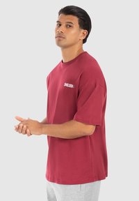 Camiseta de algodón color marrón con mangas cortas, cuello redondo y texto de logo en blanco "SMILODNX" en el pecho. Corte casual y textura suave.