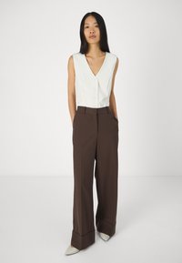 Haut sans manches blanc avec un col en V et des détails de boutons, associé à un pantalon marron à taille haute et jambe large, et des chaussures beiges à bouts pointus.