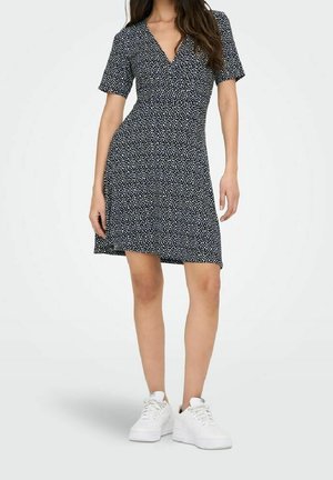 Robe portefeuille à manches courtes bleu marine à pois blancs, en tissu doux. Associée à des baskets compensées blanches. Style décontracté.