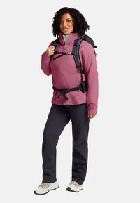 Veste rose résistante à l'eau, doublure intérieure grise, détails noirs, bretelles réglables, associée à un pantalon sombre et des chaussures blanches avec des accents noirs.