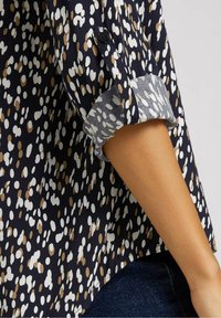 Blouse marine ornée d'un motif à pois blancs et marron clair, avec des manches retroussées et un tissu doux et léger. Coupe décontractée avec fentes sur les côtés.