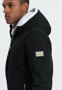 Veste à capuche noire avec doublure grise à l'intérieur, fermeture éclair et étiquette jaune accentuée sur la manche gauche. Texture lisse, design décontracté.