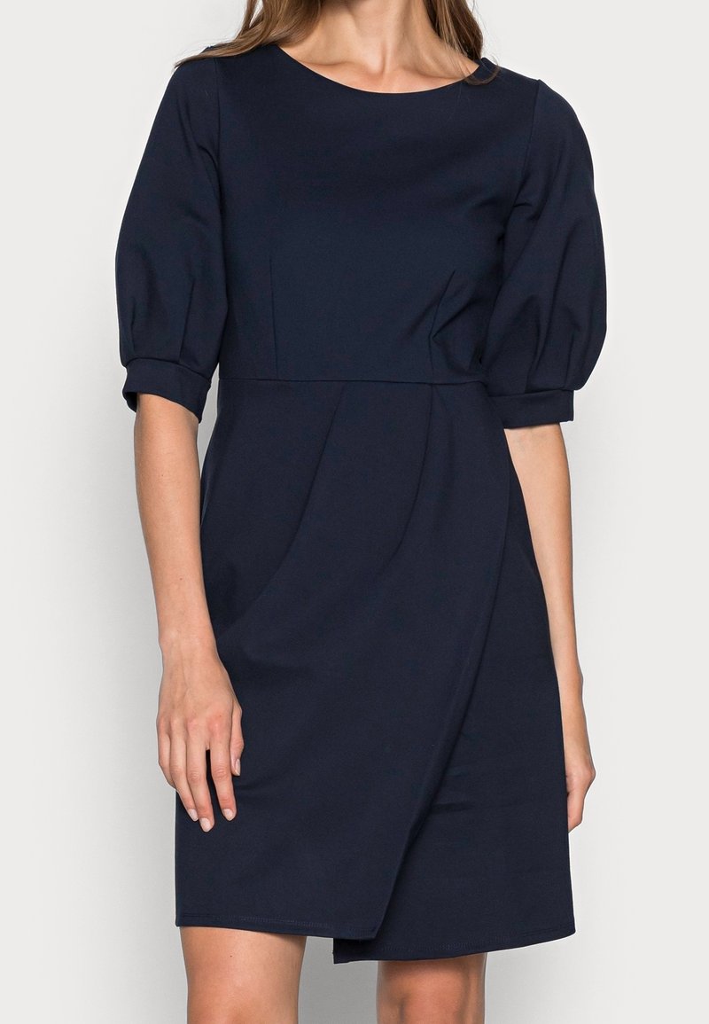 Marinblå klänning med rund halsringning och korta puffärmar. Har en figursydd bodice och detaljerad inslagning av kjolen. Slät textur.