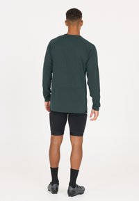 Dunkelgrünes Langarmoberteil aus strukturiertem Stoff, kombiniert mit schwarzen Shorts und Sportschuhen, von hinten gesehen.