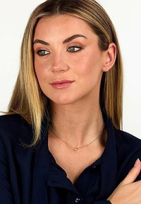 Zarte Goldkette mit herzförmigem Anhänger, getragen gegen eine dunkelblaue Bluse. Haare glatt und straff frisiert, mit einem neutralen Make-up-Look.