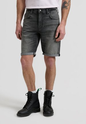 Mann trägt hochgekrempelte dunkelgraue Jeansshorts, schwarze Schnürstiefel, weiße Socken und ein weißes Hemd, steht vor einem schlichten Hintergrund.