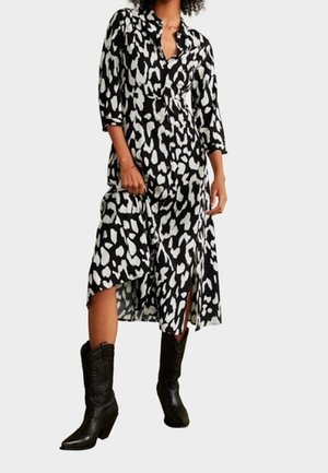 Frau, die ein schwarz-weißes Animal-Print-Midikleid mit frontalem Schlitz und schwarzen, knöchelhohen Stiefeln mit Absatz trägt, steht vor einem weißen Hintergrund.