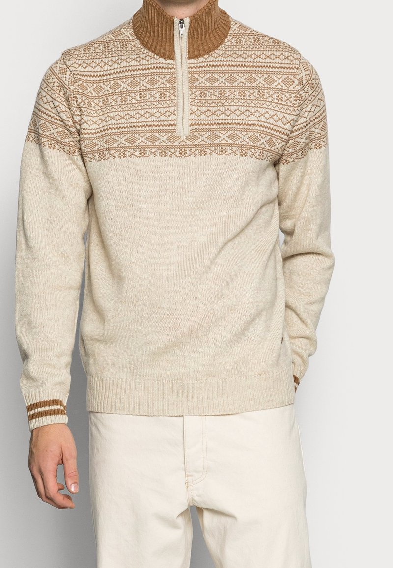 Homme portant un pull beige avec des motifs géométriques bruns sur la poitrine et les épaules, un col montant côtelé et une fermeture éclair à l'avant.