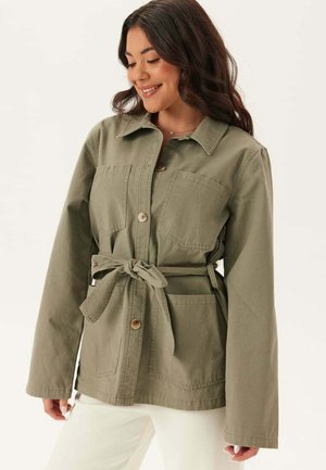 Kvinna som ler medan hon bär en olivgrön kappa med skärp och stora framfickor samt vita byxor mot en enfärgad bakgrund.