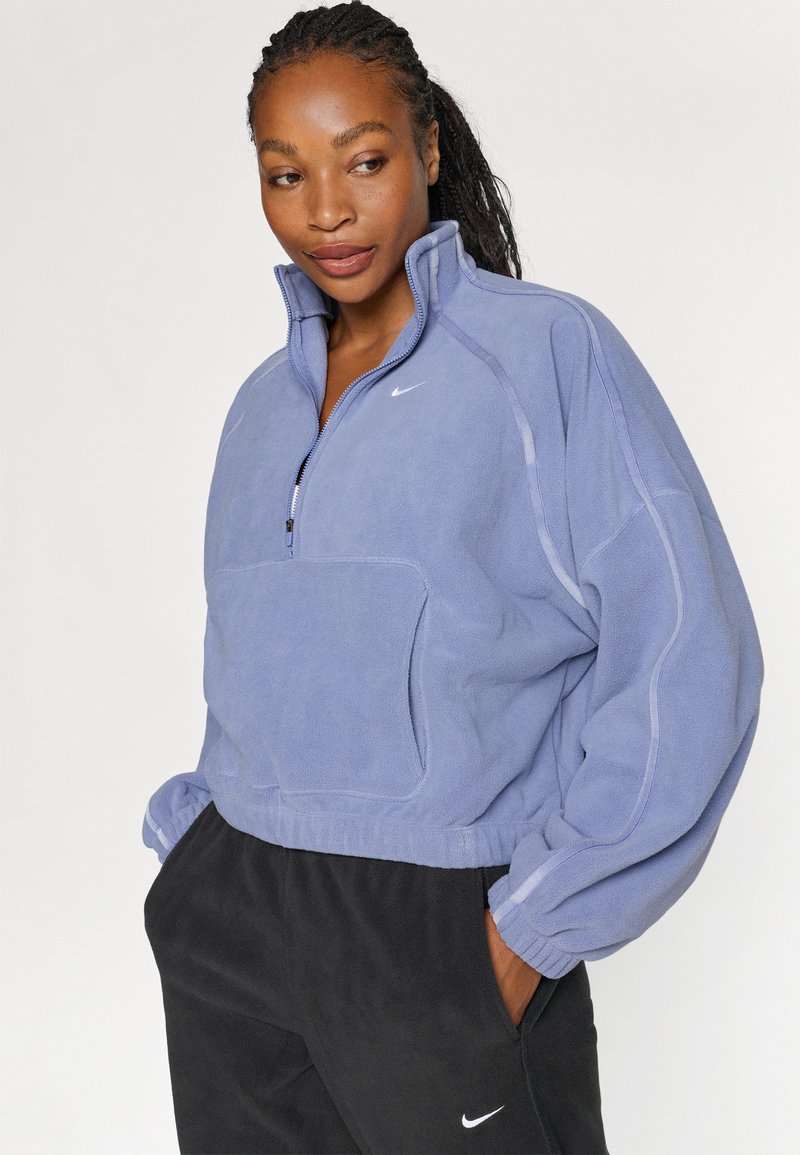 Lila Fleece-Pullover mit hohem Kragen, Viertel-Reißverschluss und Fronttasche. Mit seitlichen Paneel-Details und elastischen Bündchen. Darunter schwarze Hose.