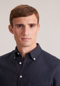 Camicia blu navy con tessuto testurizzato, taschino sul petto, bottoni bianchi e colletto. Primo piano della testa e spalle di un modello maschile.