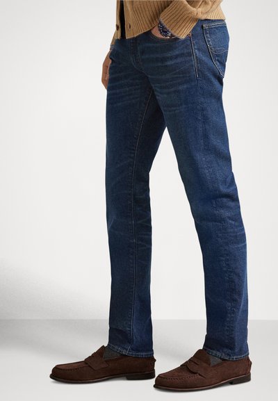 Mężczyzna w ciemnoniebieskich obcisłych dżinsach, brązowych zamszowych mokasynach i beżowym swetrze, stojący z jedną ręką w kieszeni na jednolitym tle.