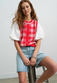Punane ja roosa ruuduline kootud vest nööpidega, koos valge puff-sleeve toppi ja heleda denim-seeliku lõhikuga.