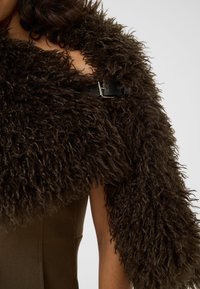 Châle en fourrure bouclée marron foncé attaché avec une sangle en cuir noire à boucle sur un vêtement olive sans manches sur le haut du corps d'une personne.