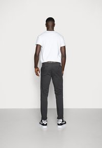 T-shirt branca de gola redonda, calças ajustadas cinza escuro e ténis preto e branco. Posição de pé mostrada de costas contra um fundo simples.