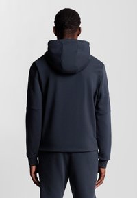 Felpa con cappuccio blu navy caratterizzata da una texture liscia, vestibilità rilassata e polsini a costine. Il design include un grande cappuccio e uno stile minimalista.