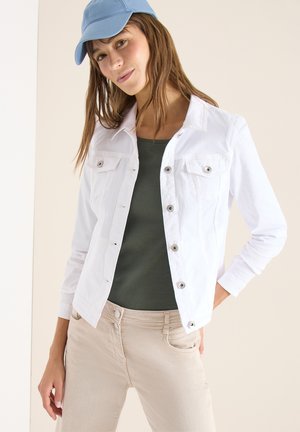 Kobieta w jasnoniebieskiej czapce, białej kurtce na guziki noszonej na ciemnozielonym topie oraz beżowych spodniach, stoi z jedną ręką w kieszeni na neutralnym tle.