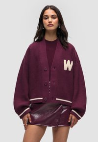 Burgundowy sweter ze splotu z dekoltem w literę V, pięcioma guzikami, paskowanymi mankietami i białą naszywką w kształcie "W" na piersi. W połączeniu z błyszcząca burgundową spódnicą.