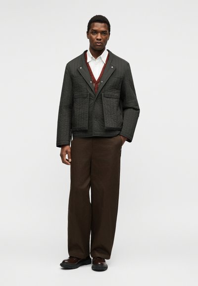 Homme debout portant une veste matelassée foncée, une chemise blanche, un gilet couleur rouille, un pantalon large marron foncé et des chaussures foncées sur fond uni.