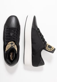 Les baskets noires présentent un mélange de matériaux en cuir et textile, un bout rond, une semelle en caoutchouc blanche et des accents de clous dorés sur la languette.