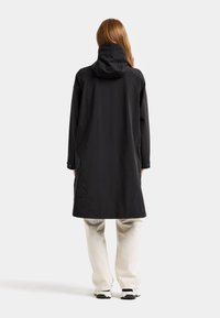 Svart vattentät jacka med huva, långa ärmar och en avslappnad passform. Har en mjuk textur och knappar vid ärmsluten. Bärs över beige byxor.