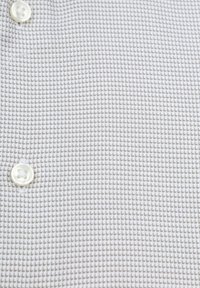 Lichtgrijze textuurstof met een klein patroon, met een witte knop op de voorgrond, wat een subtiel contrast creëert.