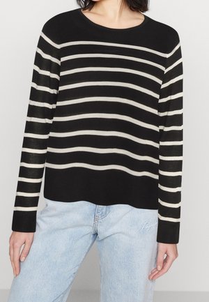 Frau trägt einen schwarzen Pullover mit weißen horizontalen Streifen und hellblauen Jeans, steht vor einem einfarbigen Hintergrund.