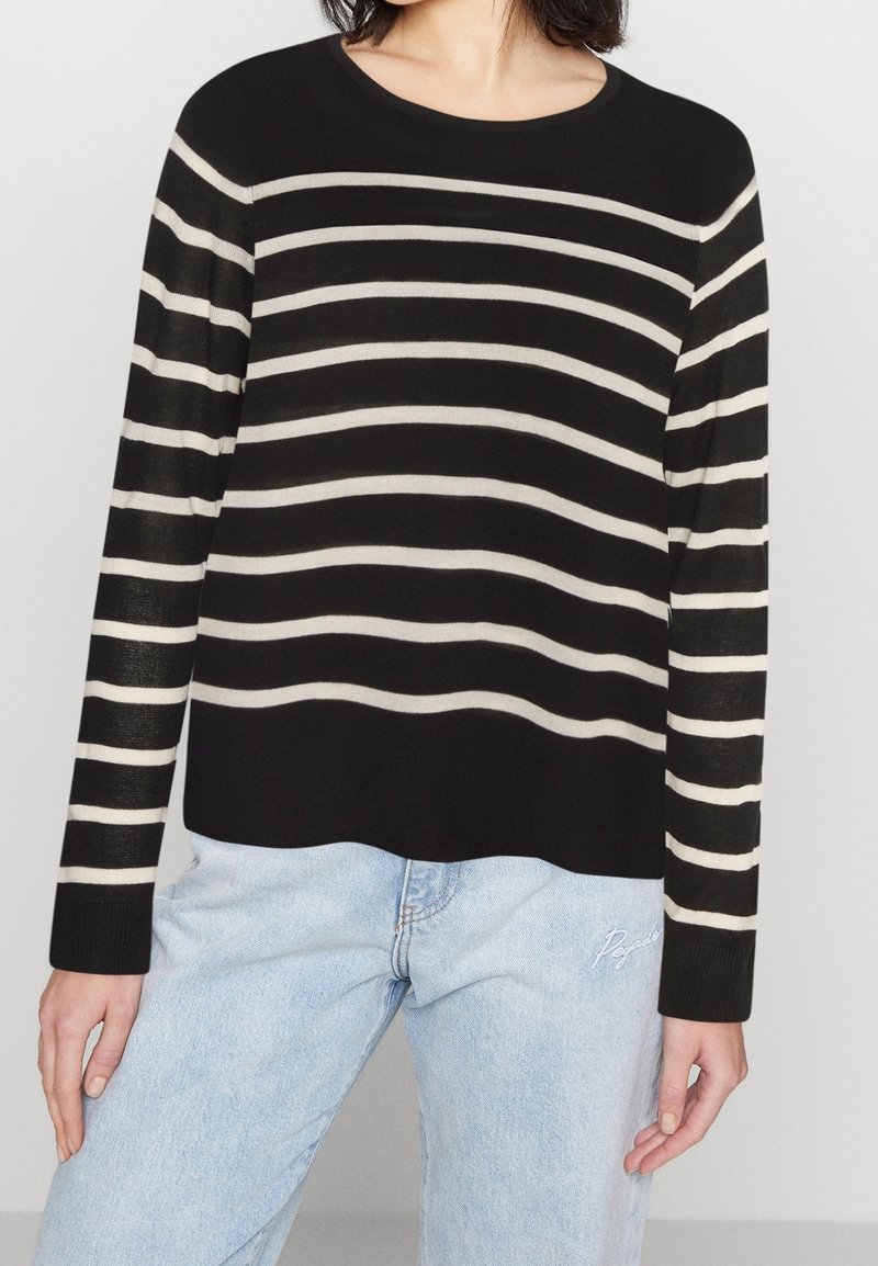 Femme portant un pull noir à rayures horizontales blanches et un jean bleu clair, debout devant un fond uni.