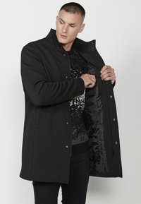Cappotto nero di media lunghezza con colletto alto, bottoni a pressione e una fodera interna liscia. Il materiale esterno è texturizzato e morbido.