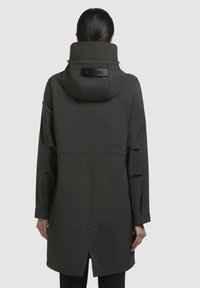 Mörkgrön vattentät jacka med huva, har hög krage, logodetalj på ryggen och en delad nederkant för ventilation. Slät tygtextur.