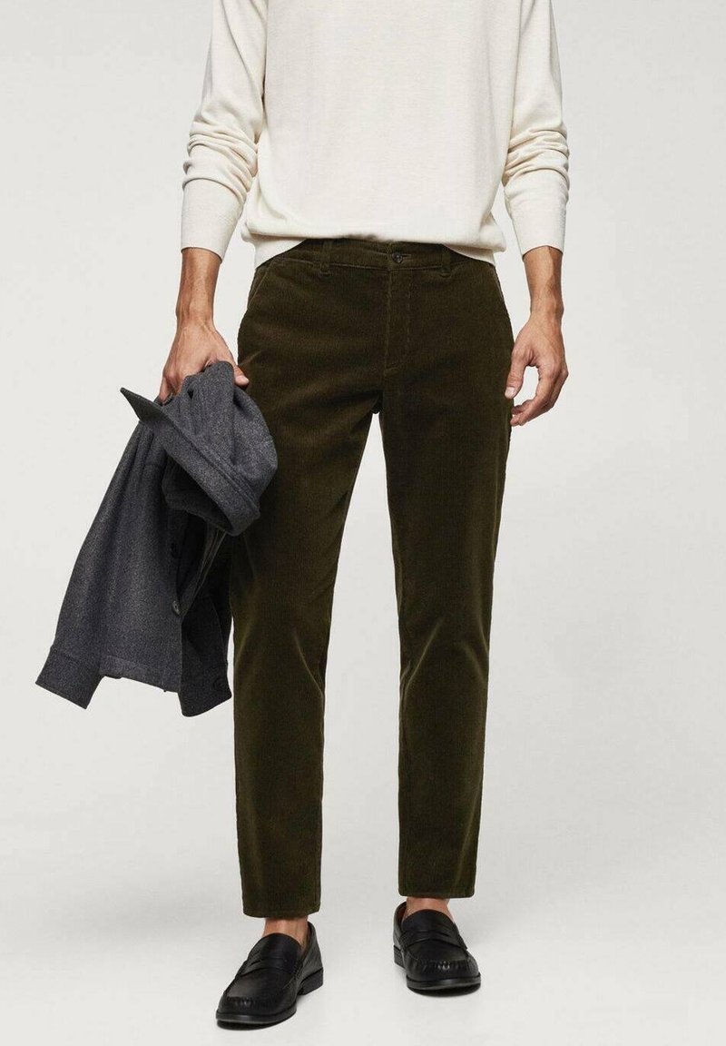 Homme portant un pantalon en velours côtelé vert olive et des mocassins noirs, tenant une veste gris foncé pliée dans la main gauche.