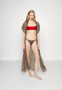 Capa longa com padrão de leopardo castanho, tecido transparente, design de frente aberta, usada sobre um top de biquíni bandeau vermelho e fio dental correspondente. Modelo de pé descalço.