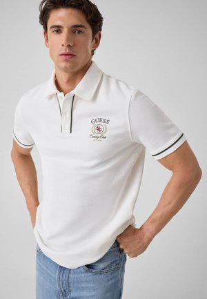 Junger Mann trägt ein weißes Guess-Poloshirt mit Logo und blaue Jeans, steht mit den Händen in der Hüfte vor einfachem Hintergrund.