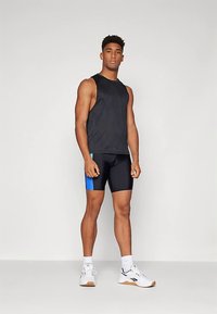 Atletski moški stoji, oblečen v črno brezrokavnik, črne in modre kolesarske kratke hlače, bele nogavice in bele tekaške čevlje na enobarvni podlagi.