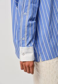 Blue striped shirt featuring white vertical stripes, a white cuff, and clear buttons. The fabric is smooth and slightly glossy.