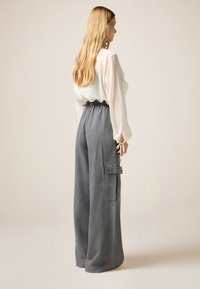Licht doorzichtige blouse met open mouwen, gecombineerd met grijze wijde pantalon met zijzakken en gestikte accenten. Effen achtergrond.